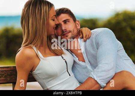 la coppia si siede vicino su una panchina, la donna con un vestito bianco e l'uomo con una camicia bianca. Lei si appoggia, sorride, mentre lui la guarda amorevolmente. Il ba Foto Stock