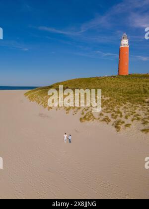 Una coppia si trova fianco a fianco su una spiaggia sabbiosa vicino ad un faro rosso e bianco, bagnata dalla calda luce del tramonto sullo sfondo dell'oceano. Foto Stock