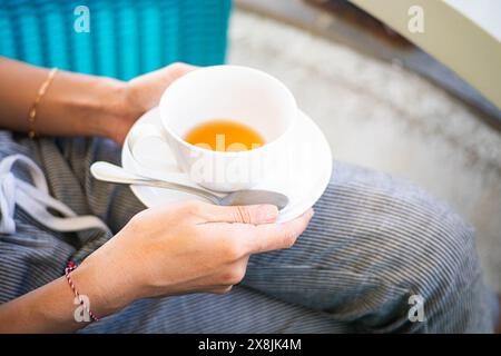 Primo piano di una persona che tiene una tazza bianca e un piattino con tè, seduta all'aperto. La persona indossa pantaloni a righe e un bracciale. Foto Stock