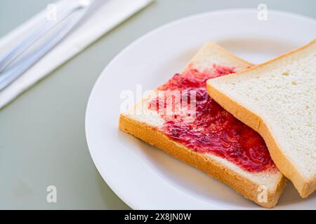 Primo piano di due fette di pane bianco su un piatto bianco, una con marmellata di fragole. Una forchetta e un coltello sono posizionati su un tovagliolo nel backgroun Foto Stock