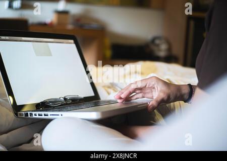 Una persona che utilizza un computer portatile su un letto con gli occhiali appoggiati sulla tastiera. Foto Stock