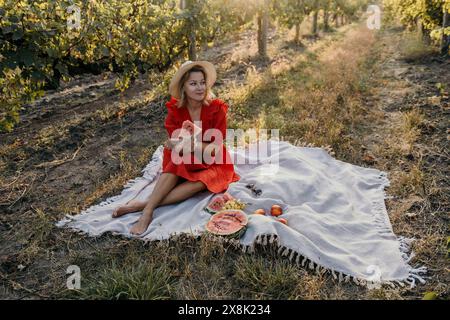 Una donna con un vestito rosso siede su una coperta in un campo, tenendo una fetta di anguria Foto Stock