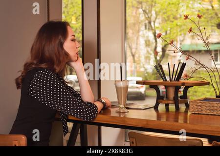 Una giovane donna elegante con un abito a pois nero è graziosamente seduta in un intimo caffè, gustando il suo caffè mentre guarda con attenzione fuori dalla finestra, l Foto Stock