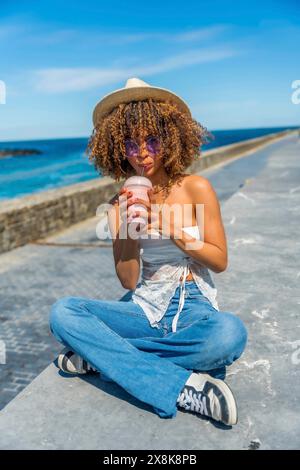 Foto verticale a tutta lunghezza di una giovane donna latina con capelli ricci sorseggiando un frullato con una paglia in riva al mare Foto Stock