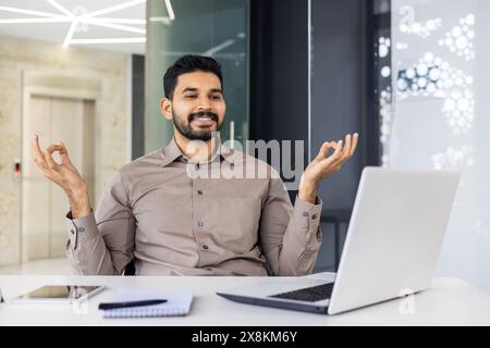 Un uomo, vestito con una camicia marrone, pratica tecniche di meditazione e rilassamento nel suo spazio di lavoro. È seduto con un notebook, un notebook e una penna, a dimostrazione dell'equilibrio in un ambiente di lavoro affollato. Foto Stock