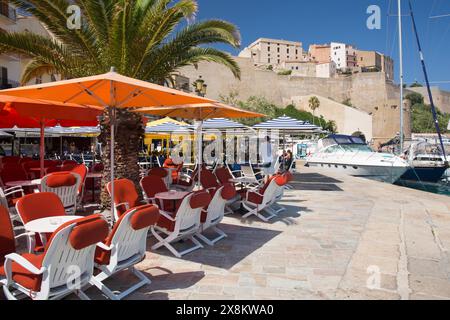 Calvi, Haute-Corse, Corsica, Francia. Colorato caffè con pavimentazione sul porto in Quai Landry sotto le massicce mura della cittadella genovese. Foto Stock