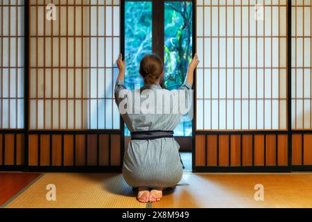 Casa tradizionale giapponese o ryokan con gaijin caucasian donna in kimono e dei tabi calzini apertura porte scorrevoli di carta shoji seduti sul tatami piano Foto Stock