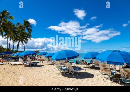 I turisti si rilassano sulle chaise longue con ombrelloni blu sulla soleggiata spiaggia di Waikiki a Honolulu, Hawaii. Foto Stock