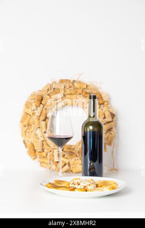 Bicchieri di vino rosso, bottiglia di vino e formaggio Brie al forno con pane tostato su un piatto bianco su un tavolo bianco con sfondo bianco e una corona di vino decorato Foto Stock