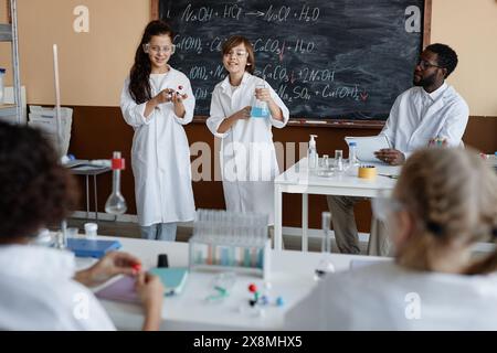 Etnie etnie diverse che indossano cappotti e occhiali da laboratorio e presentano il loro progetto davanti alla classe durante la lezione di chimica Foto Stock