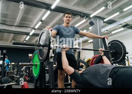 Tiro dal basso di un atleta maschile principiante che esegue una pressa da banco inclinata sotto l'occhio vigile del personal trainer in palestra. Giovane atletico Foto Stock