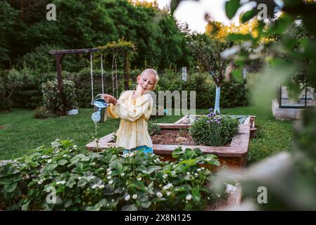 Ragazza che annaffia fragole in un letto rialzato e tiene una lattina di metallo. Prendersi cura dell'orto e piantare frutta, verdura e fiori primaverili. Foto Stock