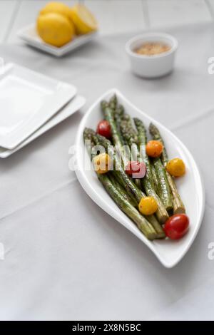 Fonte di grande fibra alimentare, asparagi verdi cotti al forno serviti in un piatto bianco, preparati come contorno con pomodori colorati e limone Foto Stock