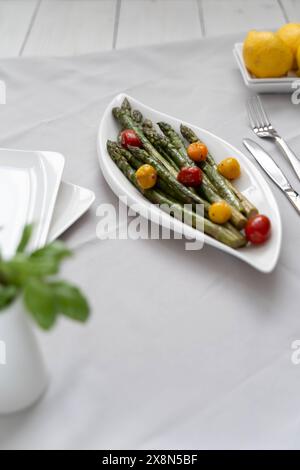 Fonte di grande fibra alimentare, asparagi verdi cotti al forno serviti in un piatto bianco, preparati come contorno con pomodori colorati e limone Foto Stock