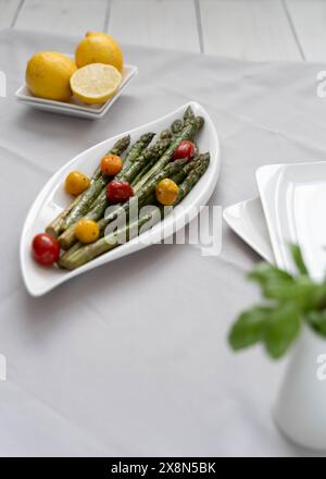 Fonte di grande fibra alimentare, asparagi verdi cotti al forno serviti in un piatto bianco, preparati come contorno con pomodori colorati e limone Foto Stock