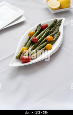 Fonte di grande fibra alimentare, asparagi verdi cotti al forno serviti in un piatto bianco, preparati come contorno con pomodori colorati e limone Foto Stock