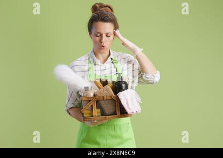 Pulizia di primavera. donna moderna di mezza età in grembiule verde e guanti in gomma isolati su sfondo verde con cestello di prodotti per la pulizia ecocompatibile. Foto Stock