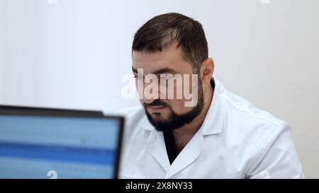 Medico di mezza età con in clinica. Fermo. Uomo barbuto in cappotto bianco che guarda il monitor del computer. Foto Stock