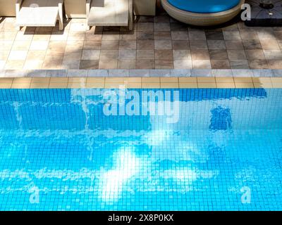 Vista dall'alto della piscina di acqua cristallina su piastrelle a mosaico a griglia blu sullo sfondo della piscina, vicino al lettino solare su pavimento in piastrelle di pietra. Spazio vuoto a bordo piscina Foto Stock