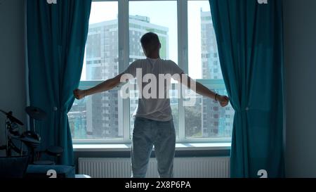 La mattina presto, silhouette dell'uomo che si allena, si allunga il corpo alla finestra a casa. Supporto. Vista posteriore di un giovane uomo che si trova contro il finestrino. Foto Stock