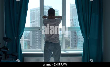 La mattina presto, silhouette dell'uomo che si allena, si allunga il corpo alla finestra a casa. Supporto. Vista posteriore di un giovane uomo che si trova contro il finestrino. Foto Stock