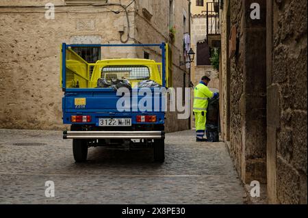 Un lavoratore municipale che raccoglie la spazzatura da una stretta strada acciottolata della città di Girona, con un camion colorato di colore blu e giallo parcheggiato. Foto Stock