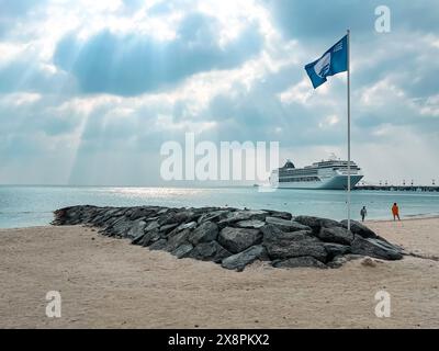 Sir Bani Yas, Emirati Arabi Uniti - 5 gennaio 2024: Una tranquilla spiaggia con una bandiera in cima a un affioramento roccioso e una nave da crociera che si crogiola tra le onde del sole che si infrangono Foto Stock