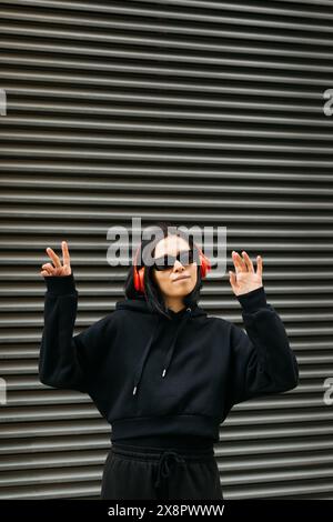Giovane donna che indossa una felpa con cappuccio nera, cuffie rosse e occhiali da sole che ballano davanti a un otturatore metallico, trasudando un'atmosfera rilassata e sicura di sé. Foto Stock