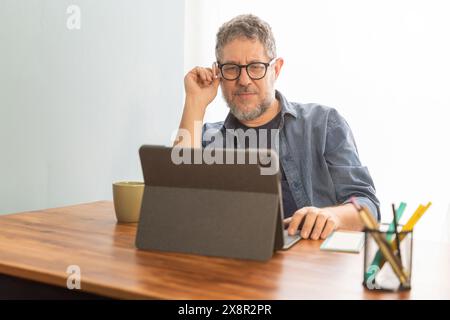 Un uomo professionista che lavora su un tablet in un ufficio domestico. La configurazione include una tazza di caffè, penne colorate e una scrivania moderna, che mette in risalto la produttività Foto Stock