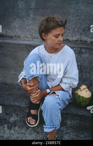 Giovane donna con i capelli corti si siede su gradini di cemento. Canggu, Bali. Foto Stock
