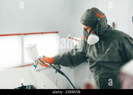 Pittore maschio in maschera respiratoria e tuta protettiva verniciatura auto con pistola a spruzzo in servizio auto Foto Stock