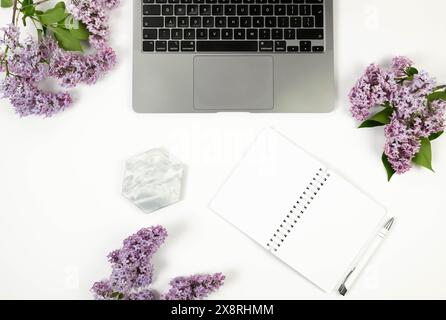 Vista dall'alto dei fiori lilla viola su sfondo bianco. Computer portatile, notebook bianco, penna e vassoio in marmo Carta da parati primaverile, piano, spazio per copie. Foto Stock