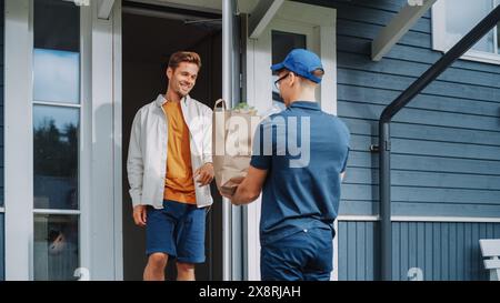 Giovane che lavora in un servizio di consegna di generi alimentari, portando un sacchetto di carta marrone pieno di verdure fresche e altri prodotti alimentari in una casa residenziale. Il destinatario felice apre la porta e firma un POD. Foto Stock