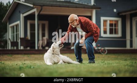 Allegro uomo anziano che si diverte all'aperto con un cane, accarezzando un giocoso White Golden Retriever. Uomo adulto felice che trascorre del tempo libero in un cortile di fronte alla casa. Foto Stock