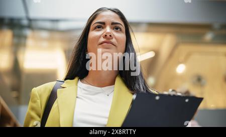 Ritratto della giovane avvocato ispanica che cammina attraverso il corridoio di un ufficio aziendale con il porta carta in mano. Agente delle risorse umane donna di successo che controlla i suoi documenti di ispezione Foto Stock