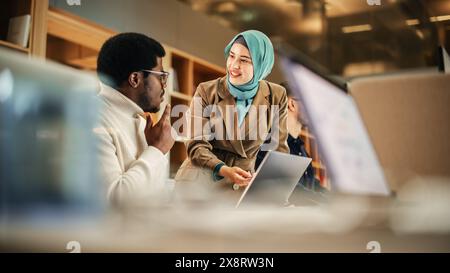 Due colleghi che collaborano e celebrano i loro risultati in un ufficio moderno. Insegnante musulmana femminile che utilizza un laptop per salire a bordo di un agente di assistenza clienti di sesso maschile nero e rispondere alla sua domanda. Foto Stock