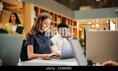 Colleghi creativi multietnici che lavorano con notebook in un ufficio affollato durante il giorno. Dipartimento risorse umane che lavora alla pianificazione. Dipendente donna sorridente Foto Stock