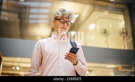 Ritratto dello studente universitario che controlla il testo sul suo smartphone e sorride mentre cammina verso l'aula. Giovane uomo bianco maschio che attraversa la sala di un grande edificio. Angolo basso, Foto Stock