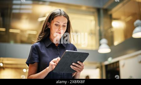 Ritratto di una potente leader donna che controlla le riunioni, che si tengono sul suo tablet e che cammina in un ufficio aziendale molto affollato. Team leader caucasico fiducioso in preparazione per una presentazione. Angolo basso Foto Stock