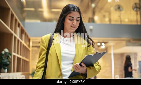 Ritratto di una donna ispanica felice che invia SMS sul suo smartphone e cammina nell'ufficio aziendale. Responsabile marketing donna che controlla le tendenze dei social media e resta aggiornato. Ripresa ad angolo basso Foto Stock