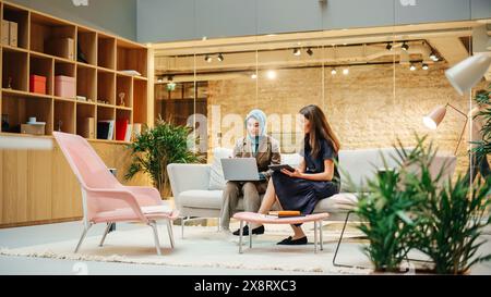 Due colleghi creativi che collaborano a un progetto di lavoro utilizzando computer e tablet in ufficio. Responsabile marketing e-commerce femminile che discute con Muslim Female Media Planner. Medio Foto Stock