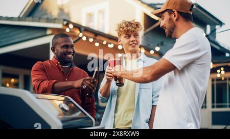 Diversi giovani adulti che trascorrono del tempo insieme a una festa all'aperto con barbecue in giardino. Amici multietnici grigliano carne alla griglia, bevono birra e chiacchierano casualmente in una calda giornata di sole. Foto Stock