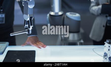 Primo piano di un moderno braccio robotico che tiene e sposta un componente meccanico in metallo. Diversi team di ingegneri che utilizzano il tablet computer per manipolare e programmare il robot. Foto Stock
