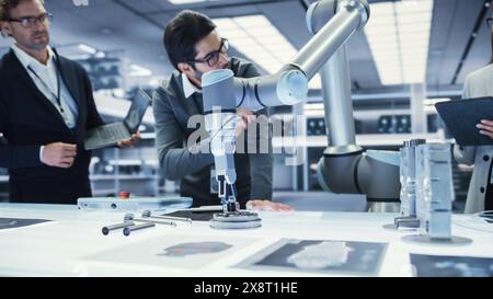 Braccio robotico moderno che tiene e sposta un componente meccanico in metallo. Diversi team di ingegneri che utilizzano il tablet computer per manipolare e programmare il robot. Foto Stock
