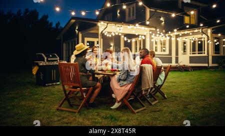 Famiglia e amici multiculturali che festeggiano fuori a casa la sera. Un gruppo di bambini, adulti e anziani si è riunito a un tavolo, facendo divertenti conversazioni. Mangiare barbecue e verdure. Foto Stock