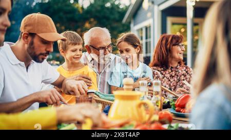 Ritratto di un nonno anziano felice che tiene i suoi nipoti brillanti e talentuosi in giro a una cena all'aperto con cibo e bevande. Famiglia che organizza un picnic insieme ai bambini. Foto Stock