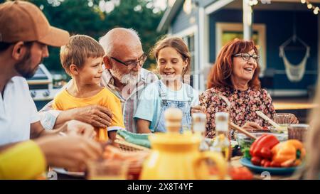 Ritratto di un nonno anziano felice che tiene i suoi nipoti brillanti e talentuosi in giro a una cena all'aperto con cibo e bevande. Famiglia che organizza un picnic insieme ai bambini. Foto Stock