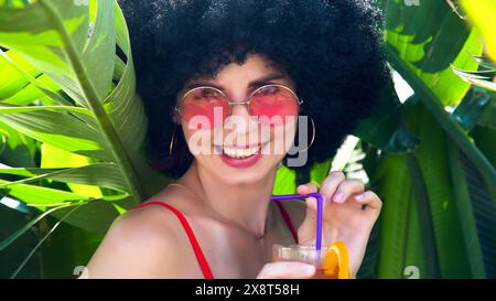 Donna tropicale in una fitta vegetazione che beve cocktail alcolici con occhiali da sole. Foto Stock