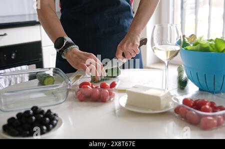 Le mani delle donne affettano cetriolo per insalata greca con Un bicchiere di vino bianco nelle vicinanze Foto Stock