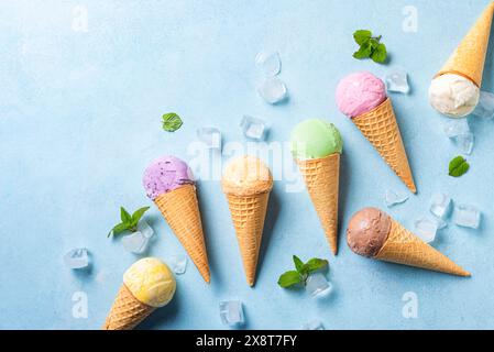 Varie palline da gelato in coni su sfondo blu, vista dall'alto, spazio per copiare. Assortimento di gelati colorati in coni per waffle con cubetti di ghiaccio, menta lea Foto Stock